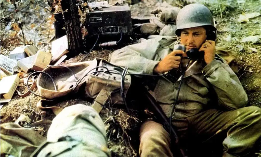 A soldier wearing a helmet uses a field radio handset in a trench, surrounded by military equipment, including a communication device and personal bags.
