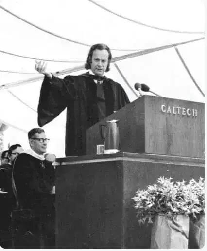 A man in academic regalia speaks at a podium labeled "Caltech" during an outdoor event. Another person in academic robes sits nearby.