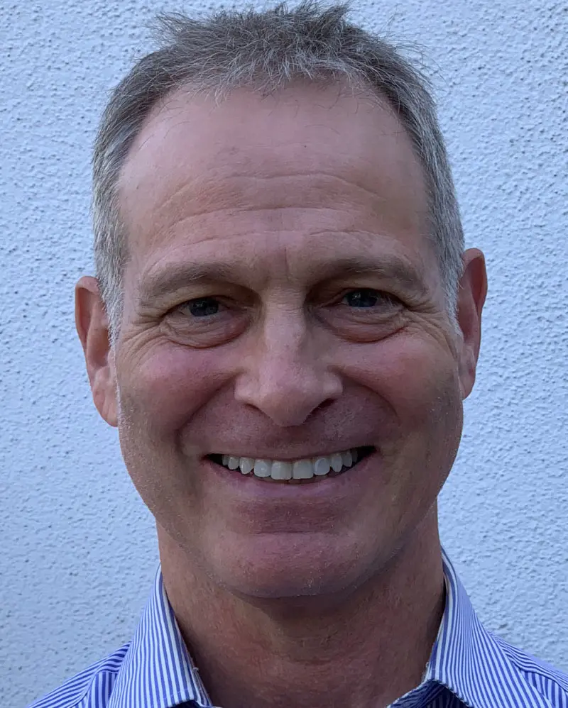 A man with short gray hair and a striped shirt smiles at the camera against a white textured background.